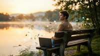 Young man sitting on park bench by lake at sunset feeding birds in peaceful golden hour nature scene.