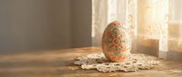 Decorative Easter egg with intricate floral patterns sits on lace doily by sunny window, creating warm spring holiday atmosphere for seasonal celebrations.