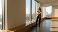 Professional businesswoman standing by floor-to-ceiling windows in modern office space overlooking city skyline, contemplating future business decisions.