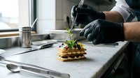 Chef in black gloves plating gourmet waffle tower with microgreens using tweezers in modern kitchen. Fine dining food preparation and plating technique.