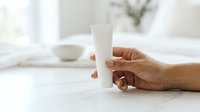 Woman with medium skin tone holding white cosmetic tube on marble surface with minimalist home decor background for skincare and beauty product marketing.