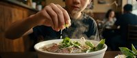 Young Asian male adding fresh herbs to a steaming bowl of noodle soup in a casual dining setting, capturing culinary preparation and food enjoyment moments.