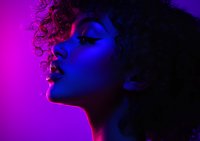 Young African American woman with curly hair in profile against dark background illuminated by vibrant neon purple and blue lighting creating dramatic contrast and mood.