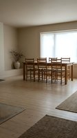 Minimalist dining room with wooden table and chairs, natural light from large window, neutral beige walls, light hardwood floors for modern home interior design.
