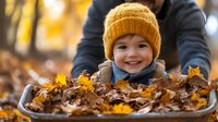 Happy Caucasian toddler boy wearing yellow knit beanie plays in pile of autumn leaves while parent holds wooden box, capturing joyful childhood moment in fall season.