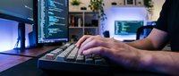 Male programmer coding at night with multiple monitors displaying code in modern home office setup with ambient lighting and plants.