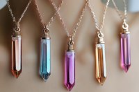 Crystal point pendants in pink, blue, purple, and amber colors hanging on delicate silver chains against soft beige background, showcasing mystical gemstone jewelry.