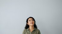 Young woman with long dark curly hair and olive skin tone wearing olive green shirt, looking upward thoughtfully against neutral gray background, suitable for motivational and aspirational concepts.