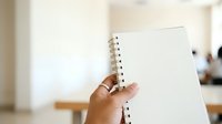 Person with medium skin tone holding open spiral-bound notebook with blank pages in bright office environment, suitable for education, planning, note-taking, and business concepts.