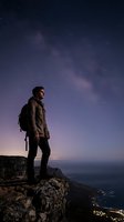 Young man with dark hair and light skin tone, wearing brown jacket and backpack, standing on rocky mountain cliff edge gazing at starry night sky with milky way galaxy visible above coastal landscape below.