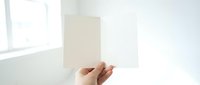 Hand holding blank white card or paper sheet in bright minimalist interior with natural window light for mockup design and branding presentation.