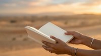 Woman with dark skin tone holding an open blank notebook against a desert landscape at sunset, suitable for education, writing, creativity, and inspirational content.