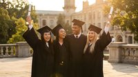 Diverse group of happy graduates in black gowns and caps celebrating with diplomas raised at university campus on sunny day.