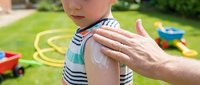 Adult applying sunscreen lotion to child's shoulder outdoors in garden during summer day for sun protection and skin care.