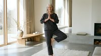 Mature woman with gray hair, Caucasian ethnicity, wearing professional black business suit, performing yoga tree pose in modern minimalist living room with large windows and contemporary furniture.