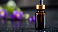 Amber glass dropper bottle with golden cap on dark surface with purple flowers and lime in blurred background for aromatherapy and essential oil products.