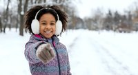 Happy African American girl with curly hair wearing winter coat and earmuffs throwing snowball in snowy park during winter season outdoor activity.