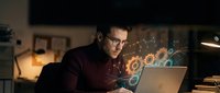 Focused businessman with glasses working late on laptop with glowing digital interface elements and data visualization in modern office environment.