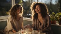 Two diverse young women with curly hair laughing together at sunset while enjoying drinks on balcony. Genuine friendship and joyful moment captured in warm evening light.