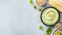Creamy avocado basil pesto sauce in black bowl with pine nuts, pasta ingredients and fresh herbs on light gray background with copy space.
