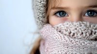 Young girl with bright blue eyes wearing cozy knitted winter hat and scarf in soft pink and beige tones, close-up portrait showing innocence and warmth.