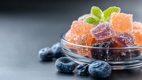 Colorful fruit jelly candies with sugar coating in glass bowl, fresh blueberries and mint leaves on dark background, selective focus with copy space.
