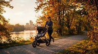 Young Caucasian mother jogging with a stroller along a scenic autumn park path at golden sunset, city skyline reflected on calm river.