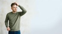 Middle-aged Caucasian man wearing olive green long-sleeve shirt and jeans, touching head with hand in casual pose against white wall background for lifestyle and fashion use.