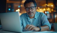 Middle-aged Hispanic businessman wearing glasses works on laptop during evening hours, illuminated by screen glow and ambient city lights in background.