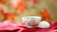 Delicate porcelain teacup with pink floral pattern and fresh white chrysanthemum flower on red silk fabric against soft autumn bokeh background.