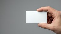 Adult male hand holding blank white business card against neutral gray background, suitable for branding, corporate identity, and marketing mockup applications.