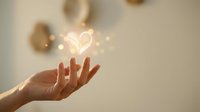Hand holding glowing heart symbol with warm light effects representing love, care, compassion and emotional wellness in soft neutral background.