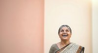 Senior woman with gray hair and glasses wearing traditional Indian saree, smiling joyfully against pink and cream colored wall background.