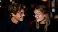 Young couple in glasses sharing intimate moment at cozy restaurant, wearing black sweaters, warm lighting creates romantic atmosphere, genuine smiles and connection.