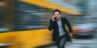 Businessman in dark coat rushing past yellow bus while talking on smartphone, conveying urban urgency and busy professional lifestyle.
