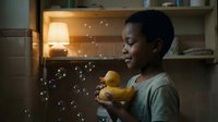 Young African American woman holding yellow rubber duck with soap bubbles floating around in warm kitchen lighting creating playful domestic scene.