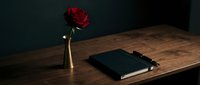 Dark moody workspace with red rose in brass vase, black notebook and pen on wooden desk. Romantic office setup for creative writing and journaling.