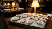 Fresh oysters on ice platter in upscale restaurant setting with warm ambient lighting and elegant table lamps creating luxurious dining atmosphere.