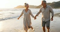 Happy mature African American couple walking hand in hand on beach at sunset, enjoying romantic vacation together during golden hour by ocean waves.