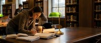 Young woman studying with books and laptop at wooden table in university library with green banker lamp and bookshelves in background for academic research.