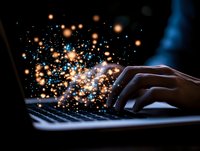 Magical glowing particles burst from laptop keyboard as person types in dark room, creating ethereal light effect with orange and blue bokeh sparkles.