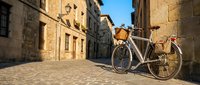 Vintage bicycle with wicker basket parked on empty cobblestone street lined with historic stone buildings under clear sky, suitable for travel and lifestyle content.