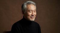 Mature Asian man with gray hair and beard in black turtleneck smiling peacefully with eyes closed against brown background for wellness and mindfulness content.