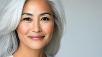 Confident mature Asian woman with silver gray hair smiling warmly at camera against neutral background for beauty and wellness campaigns.