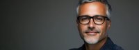 Confident mature Caucasian businessman with gray hair, beard and glasses smiling against dark background in professional studio portrait.