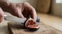 Male hand cutting fresh fig in half with knife on wooden cutting board in kitchen setting.
