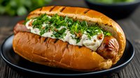 Grilled hot dog in toasted brioche bun topped with fresh chopped herbs, melted cheese and creamy sauce served on black plate against dark wooden background.