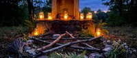 Campfire surrounded by logs at dusk with illuminated lanterns and wooden structure in forest clearing, suitable for outdoor adventure and camping concepts.