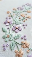 Delicate hand embroidered floral pattern with purple, green and peach flowers on white linen fabric. Traditional needlework craft for home decor and textile art.