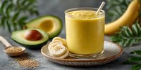 Fresh banana avocado smoothie with flaxseeds on wooden plate, surrounded by ingredients against gray background with green foliage.